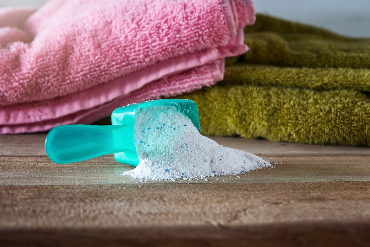 a-turquoise-scoop-of-laundry-detergent-powder-spilled-on-a-wooden-surface-in-front-of-pink-and-green-bath-towels. Laundry Detergent Powder Color Safe - Image 1